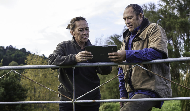 Two people on a farm leaning against a gate looking at an ipad or device